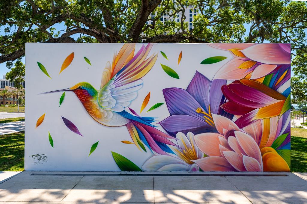 Outdoor mural of a humming bird flying near colorful flowers.