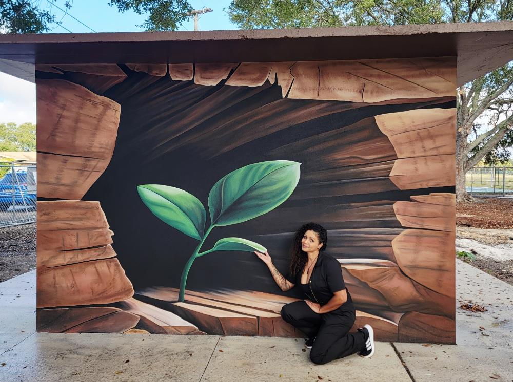 Woman kneeling in front of an outdoor mural of a large plant.